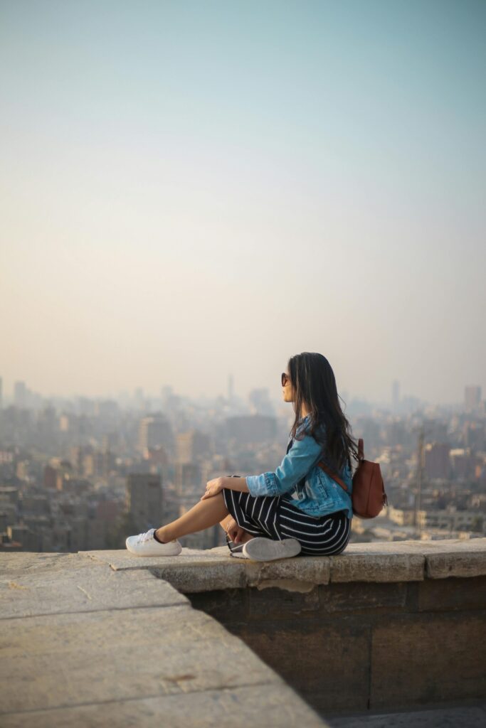 pexels-photo-1755385-1755385 A young woman in casual attire sits on a rooftop terrace, viewing a sprawling cityscape in daylight.